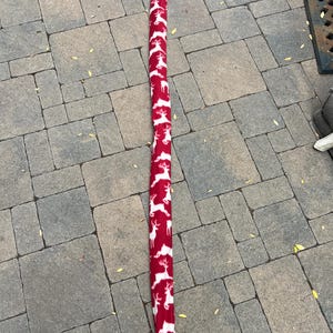 May include: A long, red fabric ski bag with a repeating white reindeer pattern. The bag is closed at the top with a black drawstring. The bag is designed to hold skis and protect them during transport.