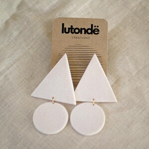May include: White geometric earrings with triangle and circle shapes, hanging from gold-colored hooks. The earrings are displayed on a brown card with the text "lutondë creations". The background is a light beige fabric.