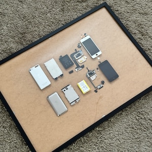 May include: A framed display of disassembled parts of a smartphone, including the back cover, screen, battery, and other components. The parts are arranged in a grid pattern on a brown background.