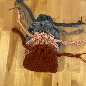 May include: Three crocheted drawstring bags stacked on top of each other. The top bag is gray, the middle bag is pink, and the bottom bag is brown.