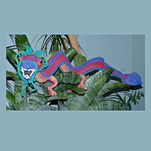 May include: A colorful crocheted dragon with blue, pink, and green accents is posed on a green leafy plant. The dragon has a detailed face with a blue mouth and orange eyes.