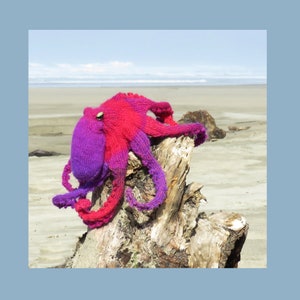 May include: A knitted octopus toy in shades of pink and purple, perched on a weathered driftwood log. The octopus has a textured, crocheted surface and is posed with its tentacles outstretched.