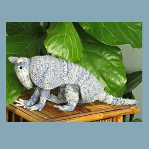 May include: A blue and white knitted armadillo toy sitting on a bamboo surface. The armadillo is posed with its head turned to the side and its legs outstretched.