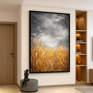 Peut inclure: Une impression d'art encadrée représente un champ d'épis de maïs secs sous un ciel nuageux et dramatique. L'œuvre est dans un cadre noir et est accrochée à un mur blanc. La palette de couleurs est dominée par des nuances de jaune, de brun et de gris.