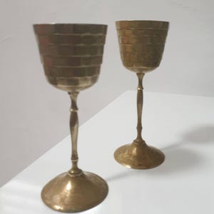 Puede incluir: Dos copas antiguas de latón con cuencos texturizados y escalonados y tallos delgados. Las copas tienen un acabado martillado y se asientan sobre una superficie blanca. El diseño general es vintage y decorativo.