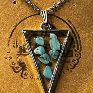 May include: Silver necklace featuring a triangular pendant. The pendant has a clear resin-like appearance with a silver border, filled with small, light blue stones. The necklace is presented on a brown card with the words "Handmade with love".