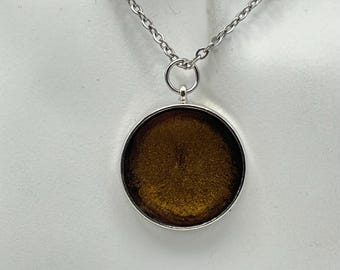 Może przedstawiać: Srebrzysty naszyjnik z okrągłą zawieszką wypełnioną głębokim, brązowym, metalicznym i mieniącym się żywicznym wkładem. Zawieszka jest zawieszona na delikatnym srebrnym metalowym łańcuszku z małymi, jednolitymi ogniwami połączonymi okrągłym uchwytem.