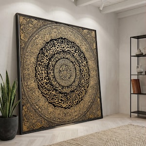 Può includere: Grande opera d'arte quadrata incorniciata con intricata calligrafia dorata su sfondo nero e oro. Il design è circolare, con il testo a spirale verso l'interno. La cornice è nera e l'opera d'arte è appoggiata a una parete bianca in una stanza con una pianta e una mensola.