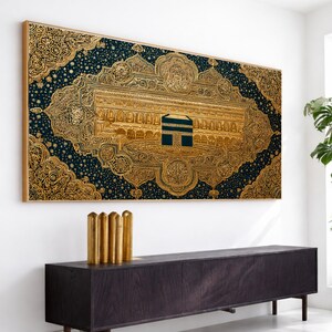 Op de afbeelding: Een ingelijst kunstwerk met een goudkleurige afbeelding van de Kaaba in Mekka, tegen een donkerblauwe achtergrond met sierlijke details. Het kunstwerk hangt boven een donkerhouten kast.