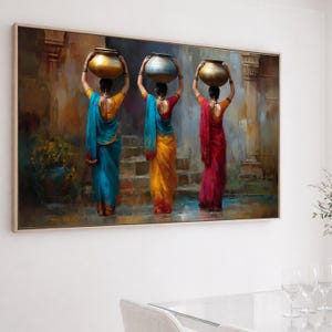 May include: A framed painting depicts three women in colorful saris, each carrying a golden or blue pot on their head. The women stand on steps, with a backdrop of architectural details in warm tones. The artwork is framed in a light-colored wooden frame.