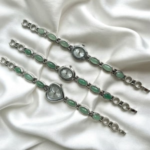 May include: Three silver-tone wristwatches with light green oval stone accents along the link bands. The watches feature different face shapes: one round, one oval, and one heart-shaped. Each watch has a white dial with silver hands and markers.