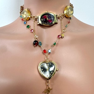 May include: A gold-toned watch choker necklace with multiple watch faces and a heart-shaped pendant. The necklace features colorful gemstone accents and a star charm. The watch faces have black and red dials.