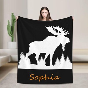 Puede incluir: Manta negra con silueta blanca de alce y diseño de bosque. El nombre "Sophia" está escrito en naranja. Una persona sostiene la manta. Ideal para la decoración del hogar.