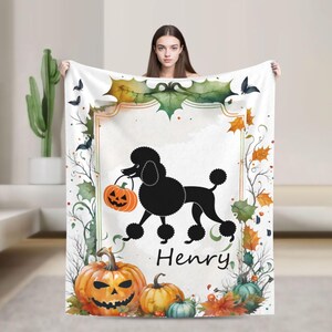 Op de afbeelding: Witte Halloween-deken met een zwarte poedel die een oranje pompoen vasthoudt. Het ontwerp bevat pompoenen, bladeren en de naam "Henry". Met decoratieve rand.