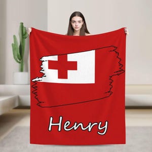 Può includere: Una coperta rossa con un disegno a croce bianca e rossa, simile a una bandiera, con il nome "Henry" in scrittura bianca. La coperta ha uno sfondo rosso pieno e un contorno nero attorno al disegno della bandiera.