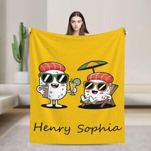 Puede incluir: Manta amarilla con un diseño de sushi de dibujos animados con gafas de sol y bebidas, con una sombrilla de playa. La manta tiene los nombres "Henry Sophia" impresos en escritura negra en la parte inferior. Los personajes de sushi son blancos con detalles rojos y verdes.