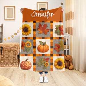 Può includere: Una coperta autunnale arancione e marrone con un design patchwork. La coperta presenta girasoli, zucche e foglie autunnali. Il nome "Jennifer" è stampato in bianco nella parte superiore.