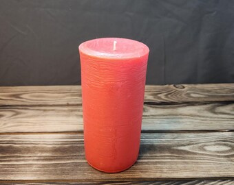May include: A single, coral-colored pillar candle with a textured surface. The candle is sitting on a wooden surface.