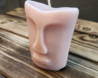 May include: A pink candle shaped like a face with a white wick. The candle is sitting on a wooden surface.