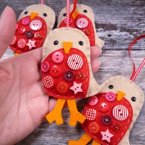 May include: Handmade felt bird ornaments with red button details. Each bird has a beige body, red breast, orange feet, and a hanging ribbon. The ornaments are decorated with various buttons and star-shaped embellishments.