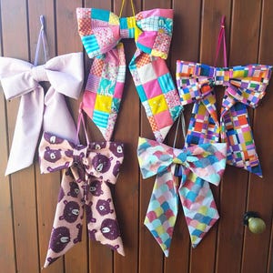 Puede incluir: Seis lazos decorativos de tela de varios colores y diseños cuelgan de una puerta de madera. Los lazos presentan diseños como patchwork, estampados de osos y formas geométricas. Cada lazo está suspendido por una cinta o cuerda.