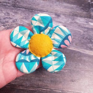 May include: A fabric flower with five petals and a yellow centre. The petals feature blue and white geometric patterns. The flower is held in a hand against a wooden background.