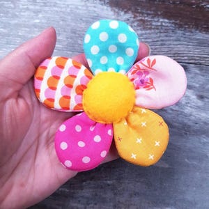 May include: A colorful fabric flower with six petals in various patterns and colors, including pink, orange, blue, and yellow. The center is a bright yellow felt ball. The flower is held in a hand.