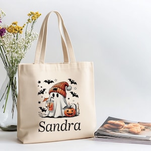 May include: Beige tote bag featuring a Halloween ghost illustration. The ghost wears an orange hat and holds a drink. Black bats and stars surround the ghost. The name "Sandra" is printed below the illustration.