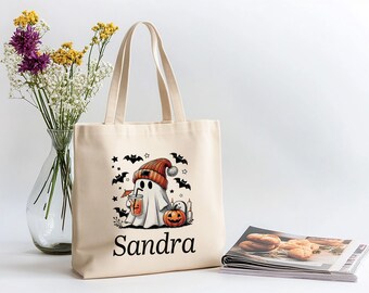May include: Beige tote bag featuring a Halloween ghost illustration. The ghost wears an orange hat and holds a drink. Black bats and stars surround the ghost. The name "Sandra" is printed below the illustration.