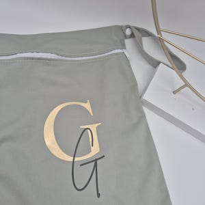 May include: A light gray zippered bag with a gold-colored "G" monogram and a black "G" script. The bag has a white zipper and a gray strap. The bag is made of a soft material.