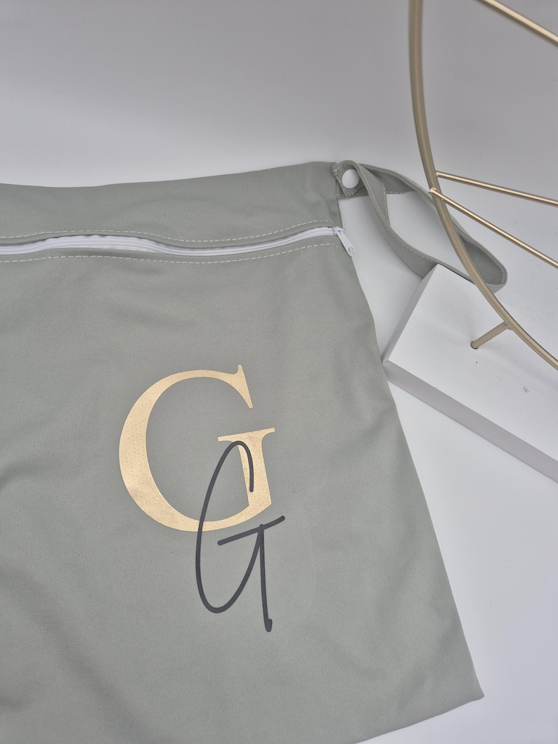May include: A light gray zippered bag with a gold-colored "G" monogram and a black "G" script. The bag has a white zipper and a gray strap. The bag is made of a soft material.