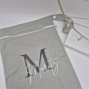 May include: A light gray zippered bag with the name "Moritz" printed on it in a stylish font. The bag has a white zipper and a small loop handle. The letter "M" is printed in a dark gray color.