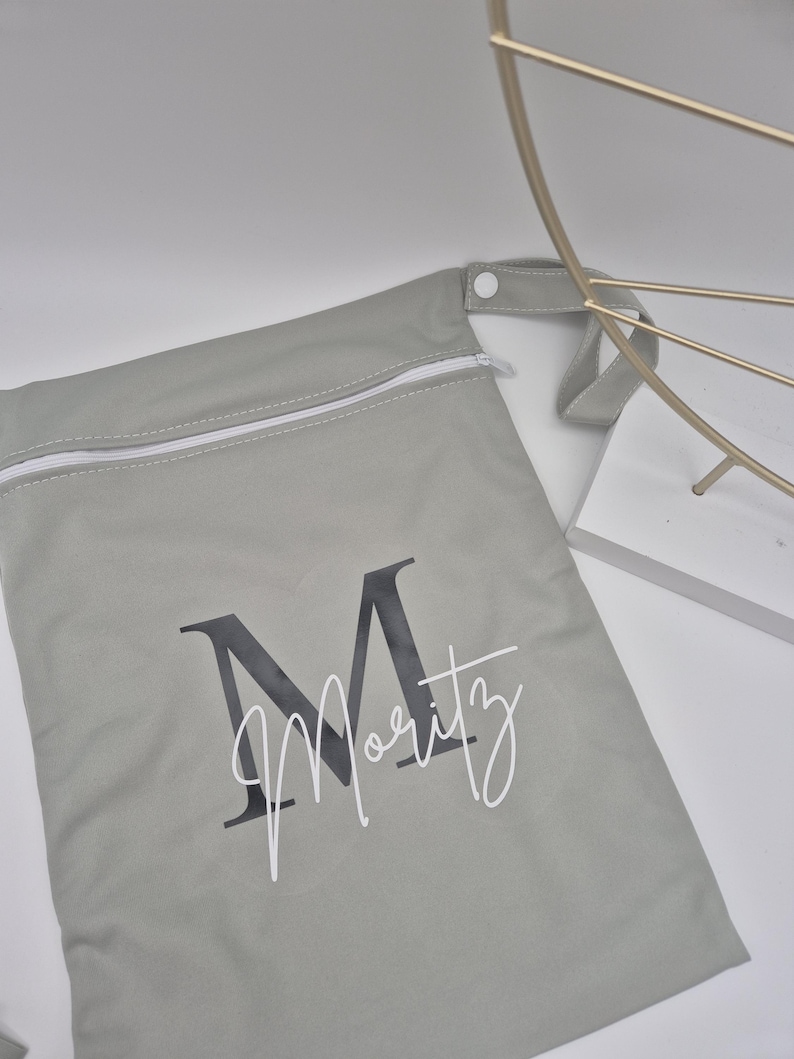 May include: A light gray zippered bag with the name "Moritz" printed on it in a stylish font. The bag has a white zipper and a small loop handle. The letter "M" is printed in a dark gray color.