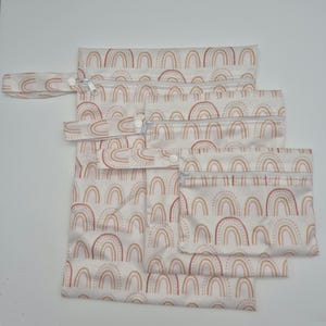 May include: A set of five reusable, waterproof bags in varying sizes. Each bag features a repeating pattern of pastel rainbows in shades of pink, peach, and brown on a white background. The bags have zippers and snap closures.