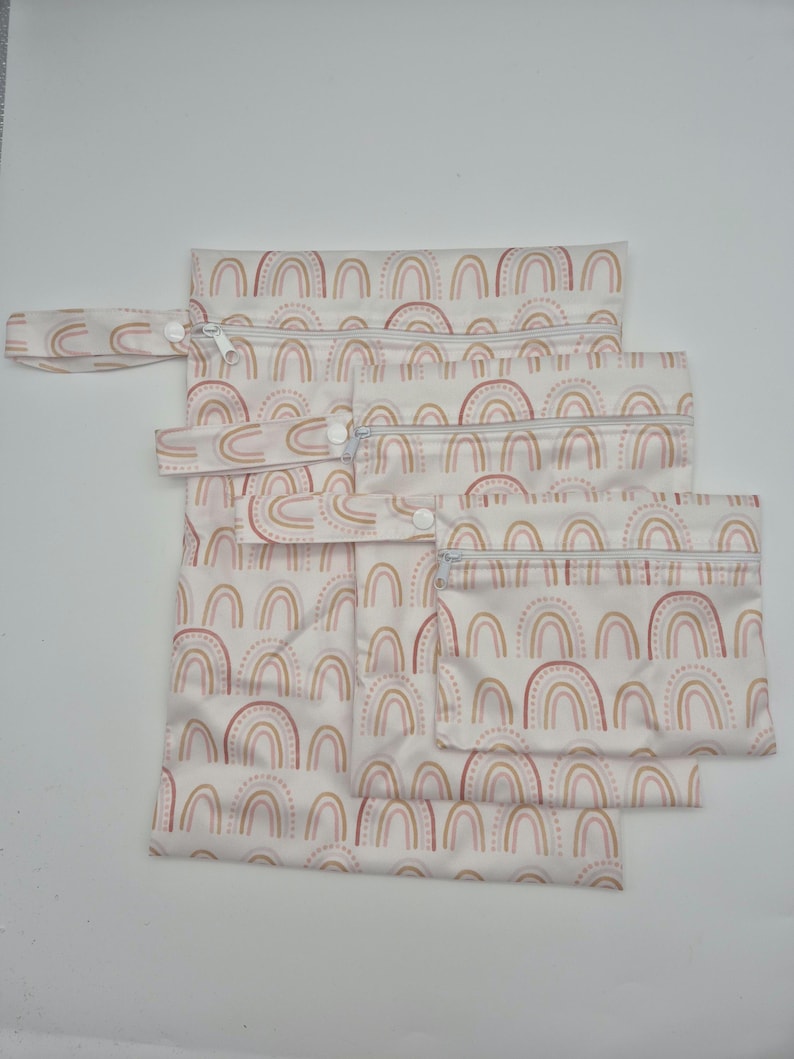 May include: A set of five reusable, waterproof bags in varying sizes. Each bag features a repeating pattern of pastel rainbows in shades of pink, peach, and brown on a white background. The bags have zippers and snap closures.