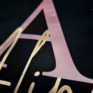 May include: Close-up of a black fabric with the name "Aline" in gold script and a large pink letter "A". The gold lettering is in a cursive font, and the pink letter is a sans-serif font.