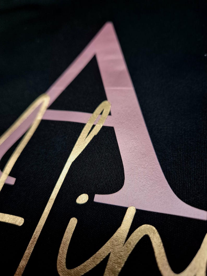 May include: Close-up of a black fabric with the name "Aline" in gold script and a large pink letter "A". The gold lettering is in a cursive font, and the pink letter is a sans-serif font.