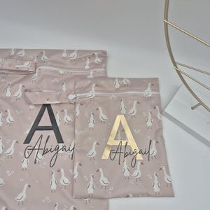 May include: Pink fabric bags with a white goose pattern and the name "Abigail" printed on them. One bag has a black "A" and the other has a gold "A". The bags have zippers.