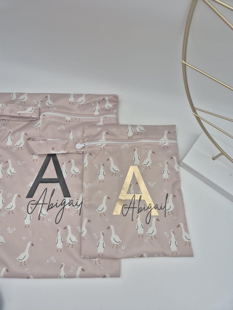 May include: Pink fabric bags with a white goose pattern and the name "Abigail" printed on them. One bag has a black "A" and the other has a gold "A". The bags have zippers.