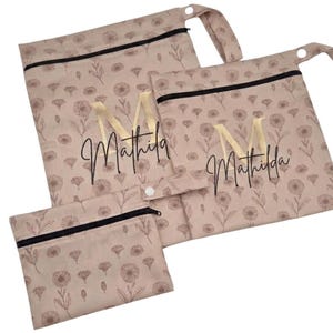 May include: A set of three beige reusable bags with a floral pattern and black zippers. The bags are personalized with the name "Mathilda" and a gold letter "M". The bags have a snap closure and a loop.