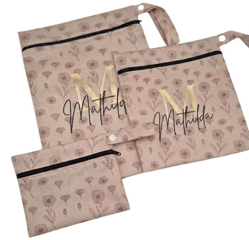 May include: A set of three beige reusable bags with a floral pattern and black zippers. The bags are personalized with the name "Mathilda" and a gold letter "M". The bags have a snap closure and a loop.