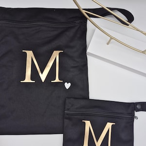 May include: Two black fabric bags with gold letter "M" and a white heart. The larger bag has a drawstring closure, and the smaller bag has a zipper. The bags are on a white surface.