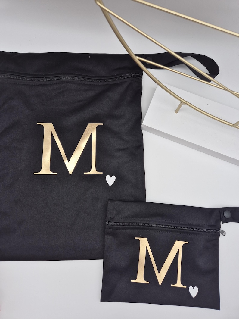 May include: Two black fabric bags with gold letter "M" and a white heart. The larger bag has a drawstring closure, and the smaller bag has a zipper. The bags are on a white surface.
