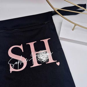 May include: Black zippered pouch with the letters "SH" in pink, and the name "Sophie Hingley" in a cursive font. A small pink heart is visible. A gold-colored decorative object is in the background.
