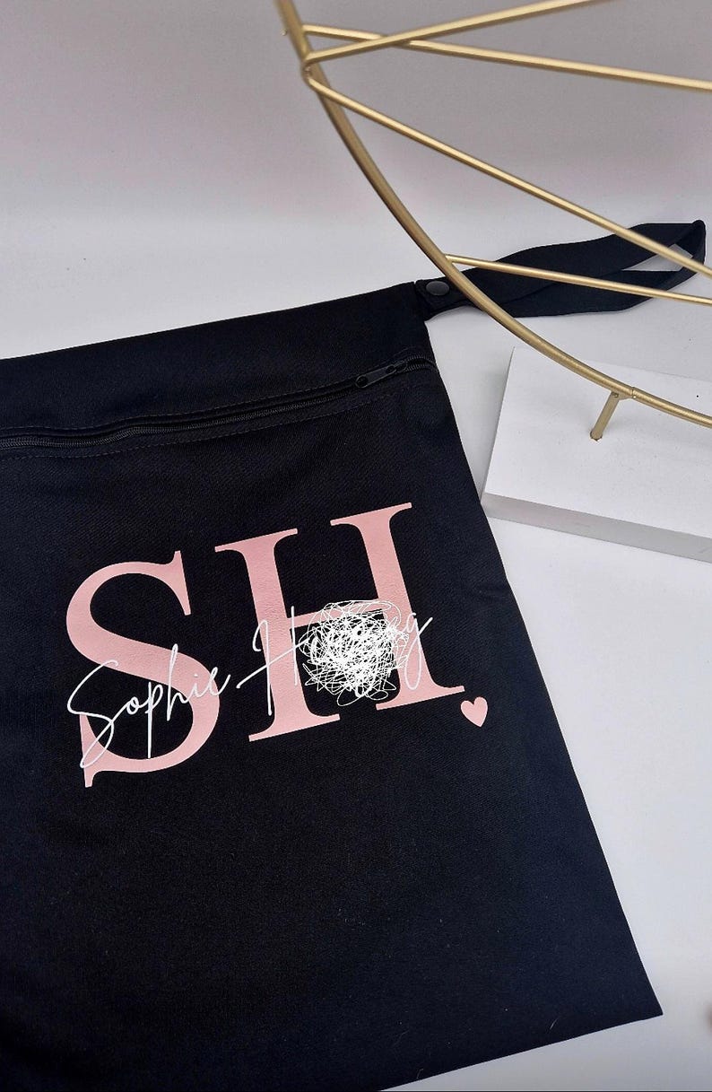 May include: Black zippered pouch with the letters "SH" in pink, and the name "Sophie Hingley" in a cursive font. A small pink heart is visible. A gold-colored decorative object is in the background.