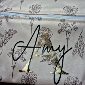 May include: A beige floral-patterned pouch with a white zipper. The pouch features the name "Amy" in a black script font with a gold foil letter "A". The floral design includes various flower and leaf illustrations in a brown color.