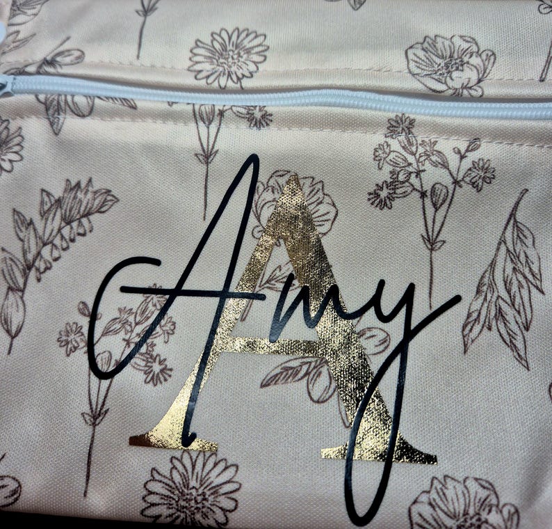 May include: A beige floral-patterned pouch with a white zipper. The pouch features the name "Amy" in a black script font with a gold foil letter "A". The floral design includes various flower and leaf illustrations in a brown color.