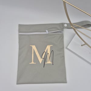 May include: A gray zippered pouch with a white zipper and a handle. The pouch features the letter "M" in gold with a handwritten "m" underneath. The pouch is likely for storage or carrying small items.