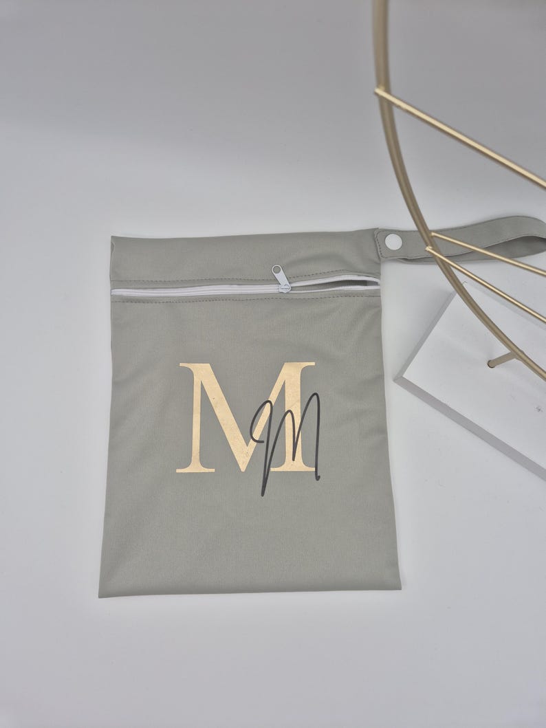 May include: A gray zippered pouch with a white zipper and a handle. The pouch features the letter "M" in gold with a handwritten "m" underneath. The pouch is likely for storage or carrying small items.