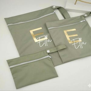 May include: Three sage green zippered pouches of varying sizes. The largest has a handle. Each pouch features the name "Ella" in a gold letter "E" and script font. The zippers are white.